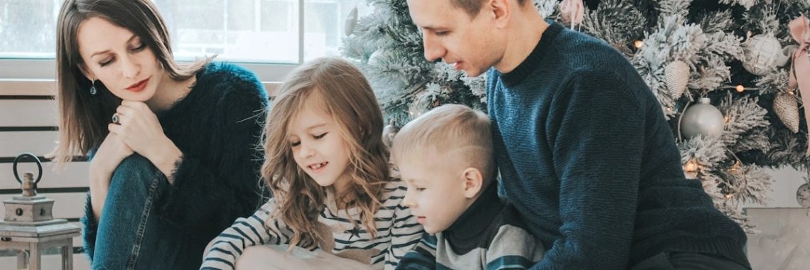 girl and boy reading book sitting between man and woman beside Christmas tree