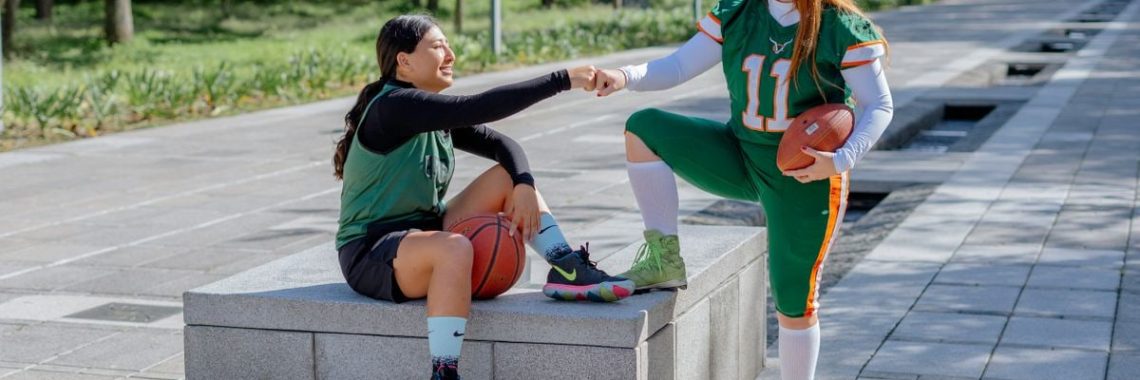 Two female athletes share a fist bump while sitting outdoors, holding sports equipment in Mexico City.